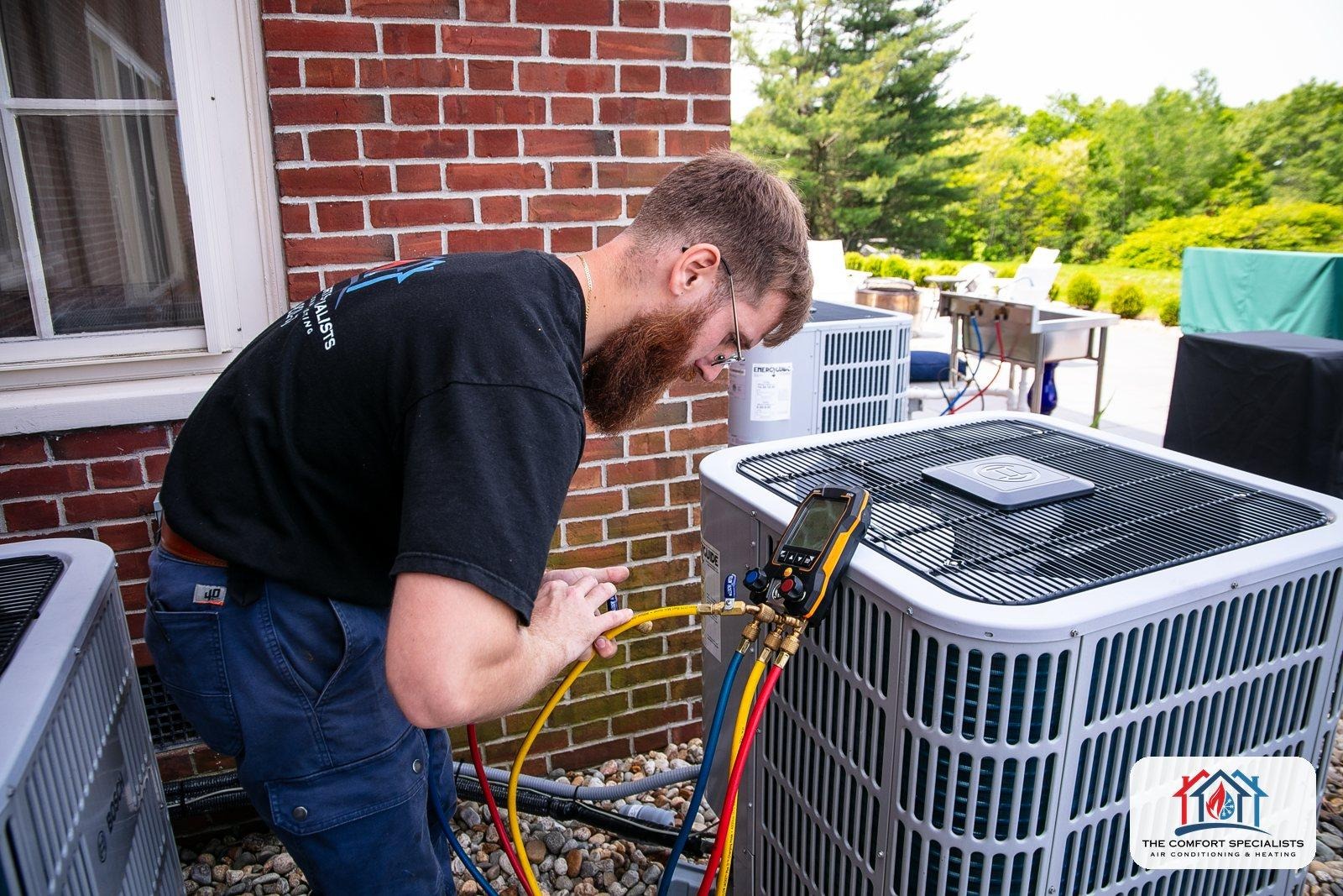 A technician checks an outdoor heat pump with gauges beside a brick home in Central Massachusetts A technician checks an outdoor heat pump with gauges beside a brick home in Central Massachusetts